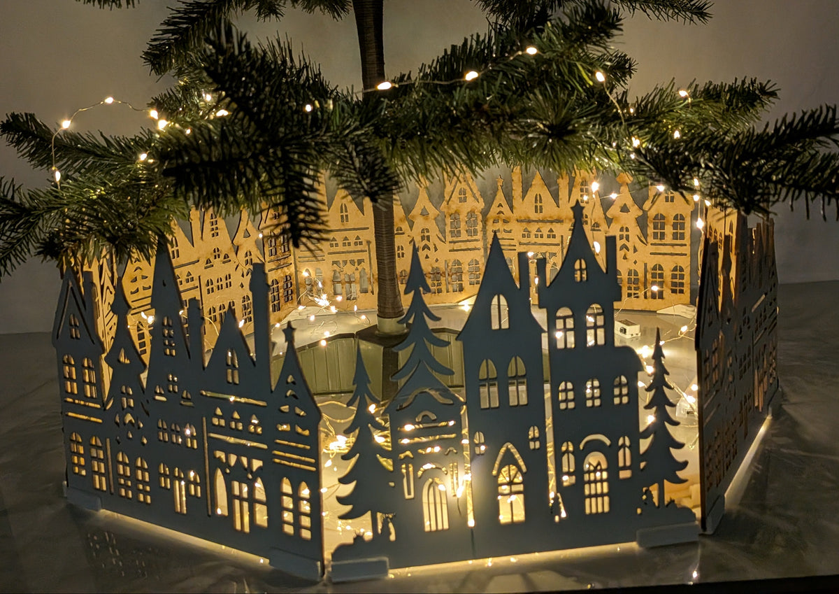A hexagonal LED light box featuring a silhouette cutout of old European townhouses and trees, glowing with warm yellow lights, styled under a Christmas tree with fairy lights.