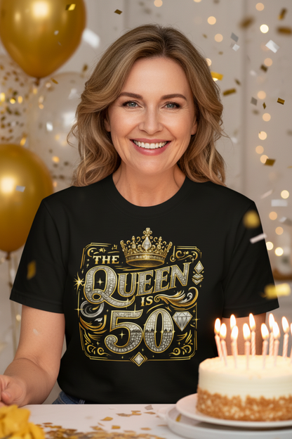 A close-up of a person wearing a black short-sleeve T-shirt with a large decorative graphic on the chest reading 'THE QUEEN IS 50' in gold, silver, and white embellished lettering with a crown and diamond motifs.