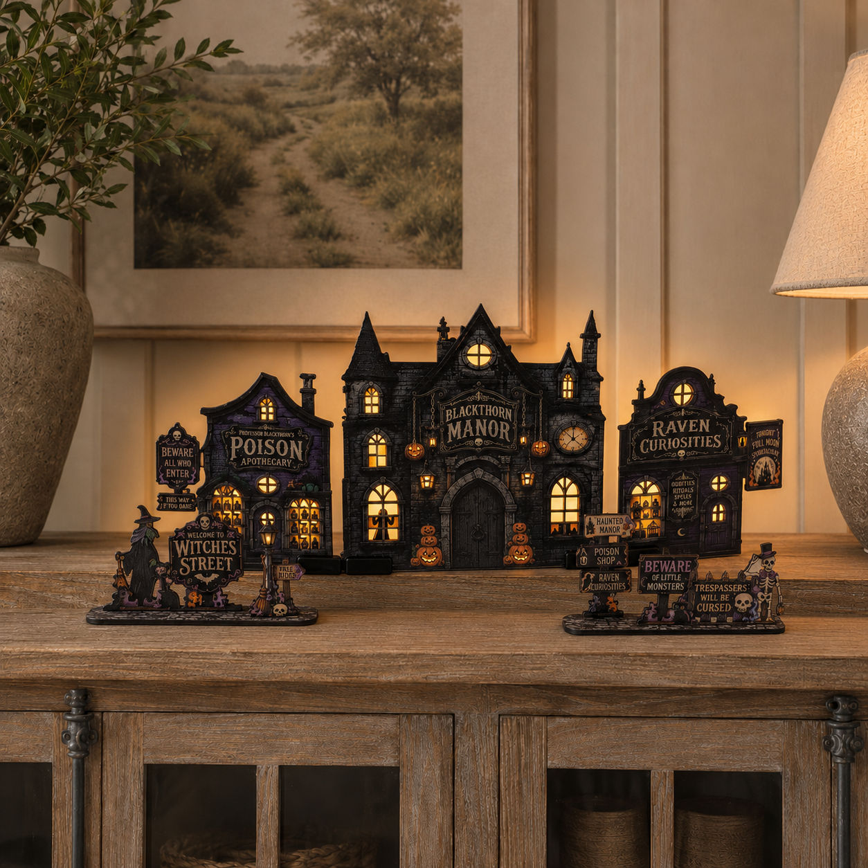 A set of Halloween-themed miniature decorative buildings and signs displayed on a wooden cabinet in a warmly lit room. The collection includes a central haunted manor labeled 'Blackwood Manor' with glowing windows, flanked by smaller spooky structures such as 'Poison Wonderland,' 'Witches Street,' 'Raven Colossus,' and 'Cemetery Gates.