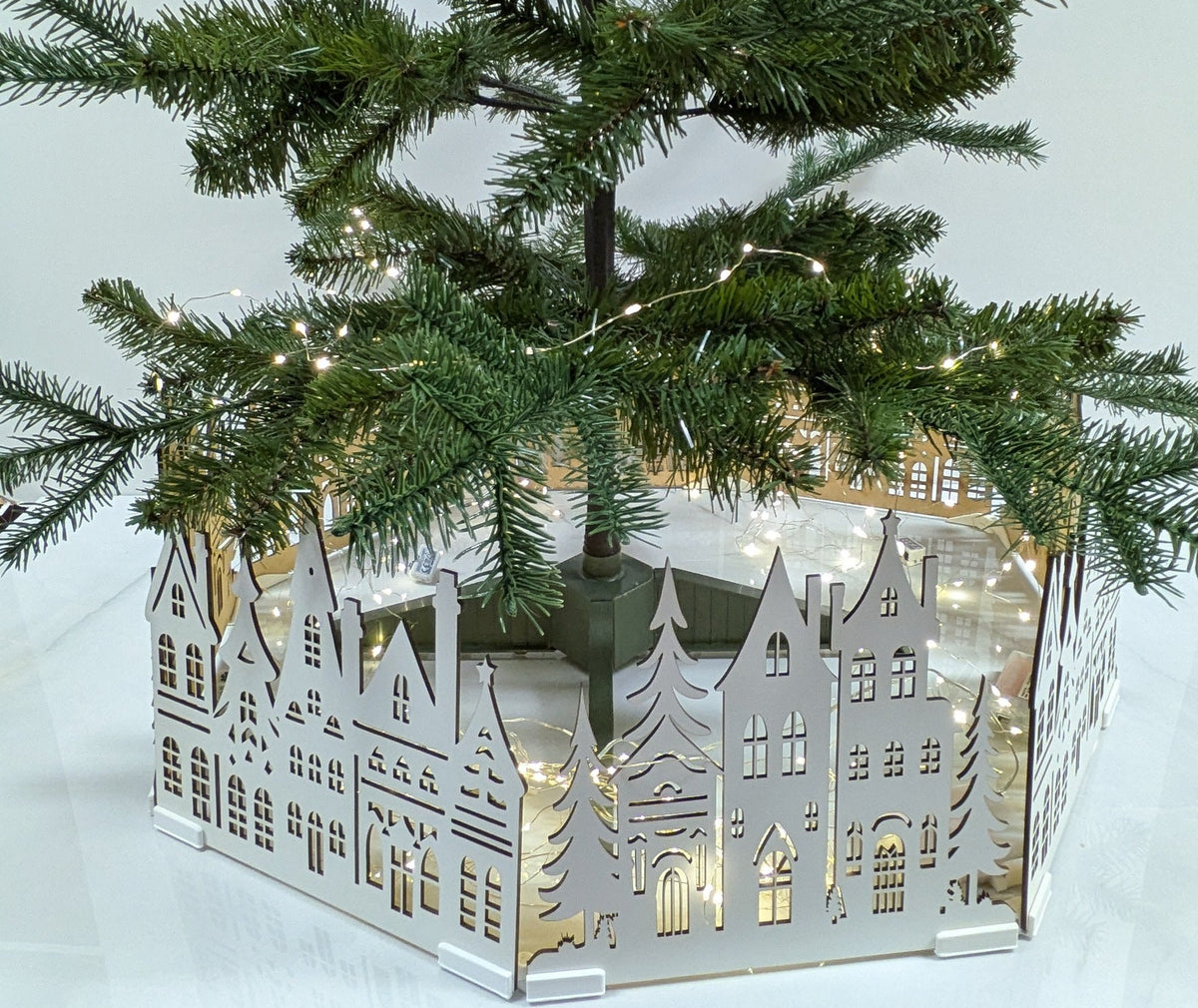 A small artificial Christmas tree with green needles and warm white string lights, planted in a white hexagonal decorative base featuring a cutout village scene, set on a white tabletop with a wooden drawer cabinet below.