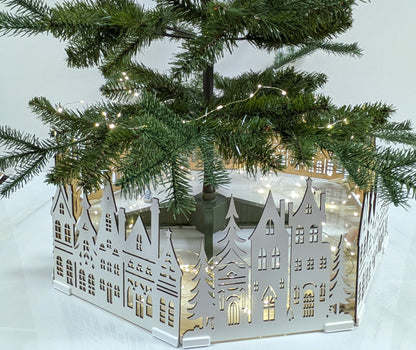 A small artificial Christmas tree with green needles and warm white string lights, planted in a white hexagonal decorative base featuring a cutout village scene, set on a white tabletop with a wooden drawer cabinet below.