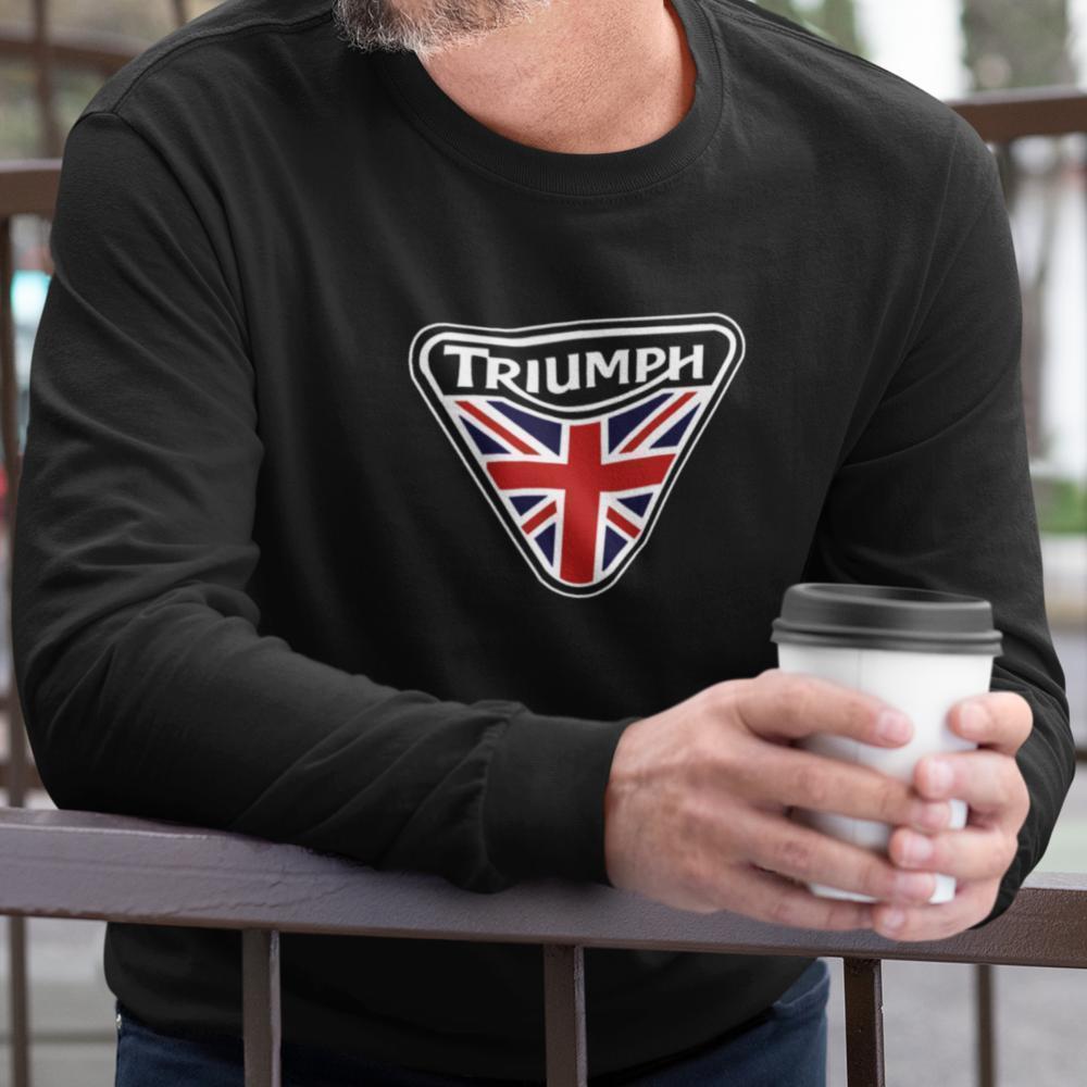 A man wearing a black long-sleeve shirt with a Triumph motorcycle logo featuring the Union Jack flag on the chest, holding a white coffee cup while leaning on a railing.