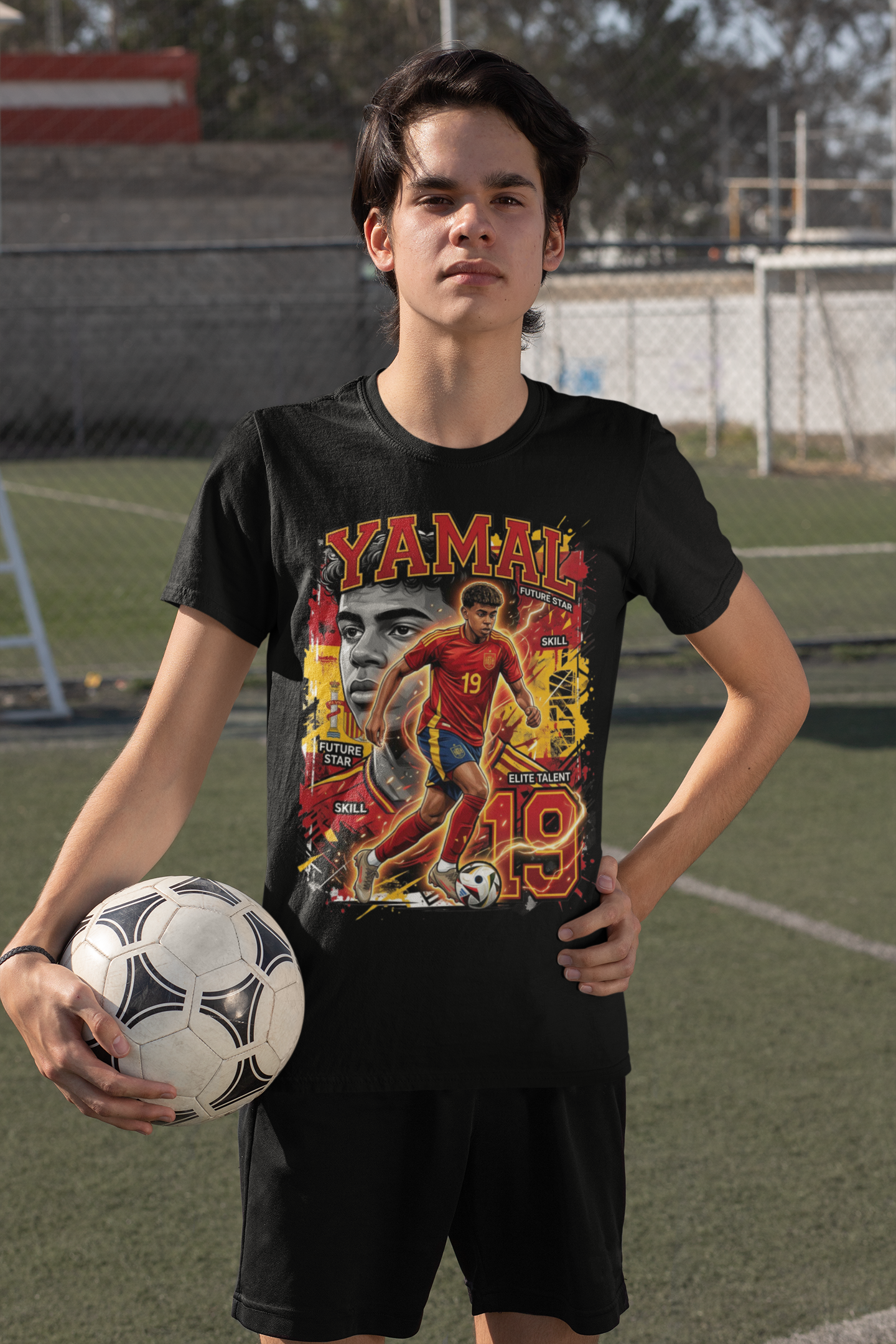 A young man wearing a black graphic t-shirt with a vibrant soccer design featuring the name 'Yamal' and the number 19, holding a soccer ball on an outdoor turf field.