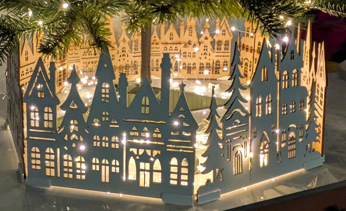 A decorative white wooden box with a silhouette cutout of old-style houses and trees, filled with warm white fairy lights, placed on a tabletop beneath a real Christmas tree.