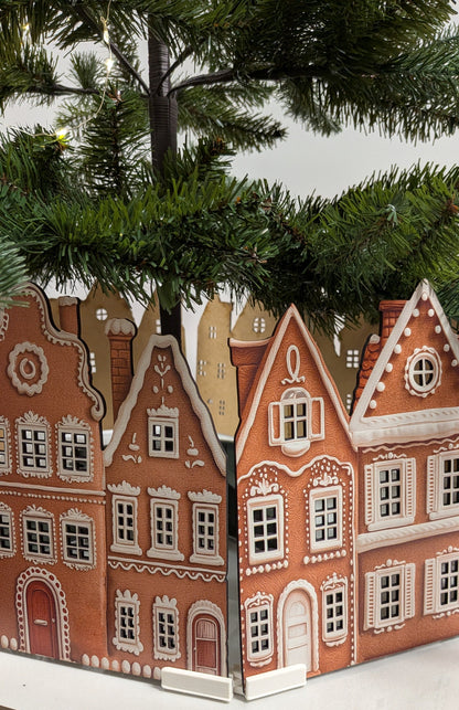 A decorative miniature village scene with orange-brown traditional house facades printed on a card or panel, arranged on a white surface with evergreen branches above.