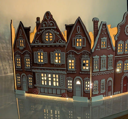 A decorative miniature model of traditional European townhouses with illuminated windows, displayed on a reflective surface against a plain beige background.