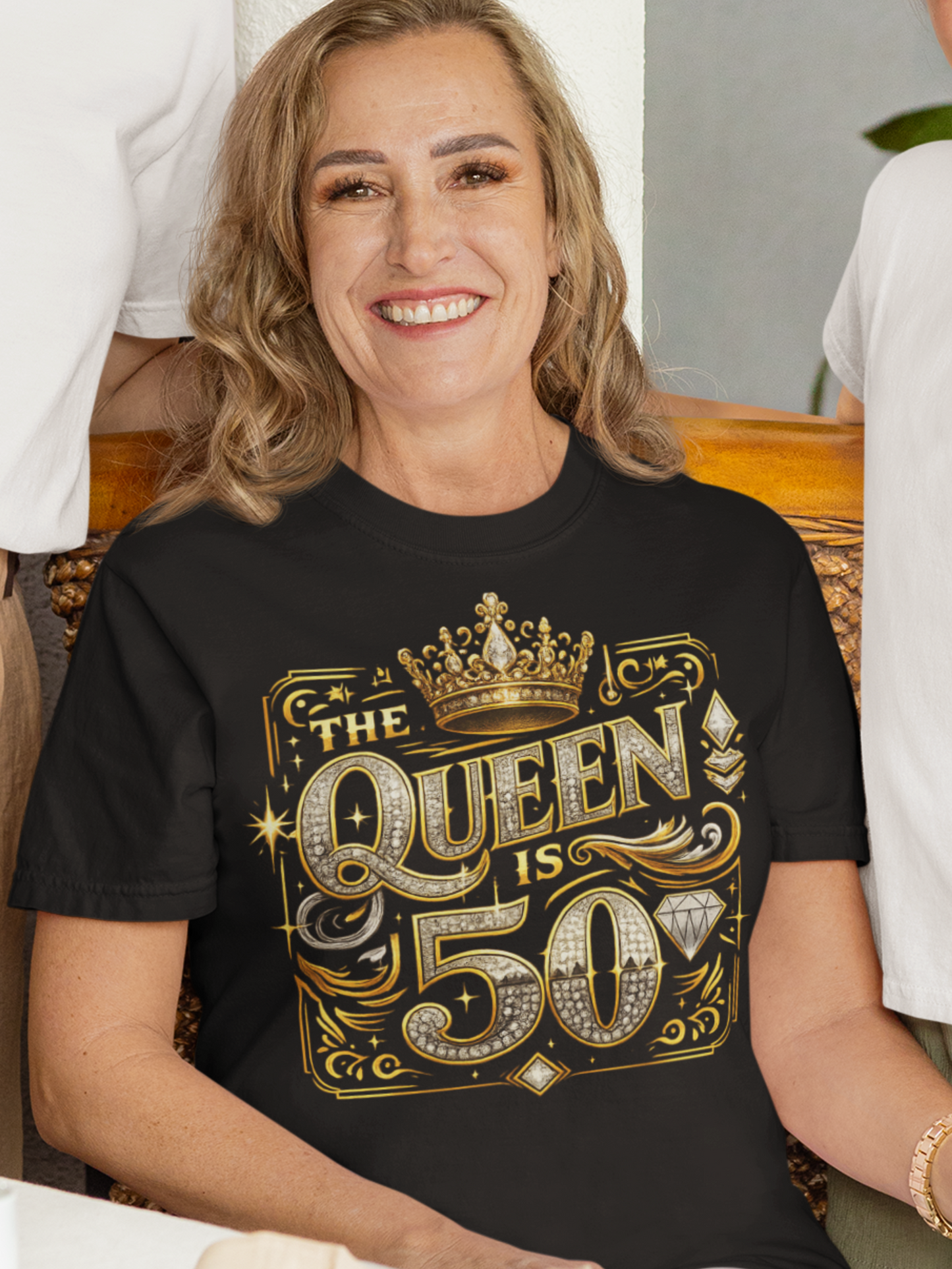 A smiling woman wearing a black short-sleeve t-shirt with a large gold and white graphic that reads 'THE QUEEN IS 50' with a crown and diamond motifs.