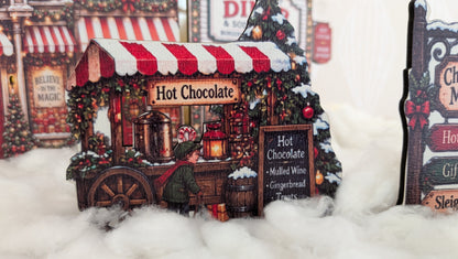 A miniature hot chocolate stand decoration with red and white striped awning, snow-dusted details, and a festive Christmas tree beside it, displayed in a snowy decorative setting.
