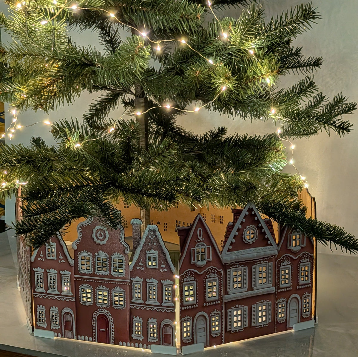 A decorative Christmas tree trunk box shaped like a row of red and beige gabled houses, illuminated from within, with a real Christmas tree and warm string lights draped over it on a tabletop.