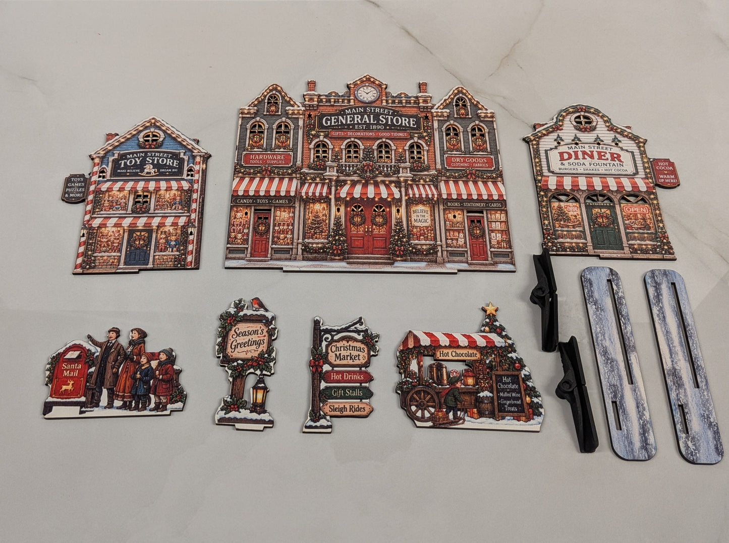 A set of holiday village-themed accessories arranged on a light marble surface. The set includes three storefront magnets (General Store, Diner, and Tavern), four character/sign magnets, two black plastic handles, and two silver/gray rectangular pieces, with fairy lights visible in the background.