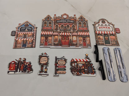 A set of holiday village-themed accessories arranged on a light marble surface. The set includes three storefront magnets (General Store, Diner, and Tavern), four character/sign magnets, two black plastic handles, and two silver/gray rectangular pieces, with fairy lights visible in the background.