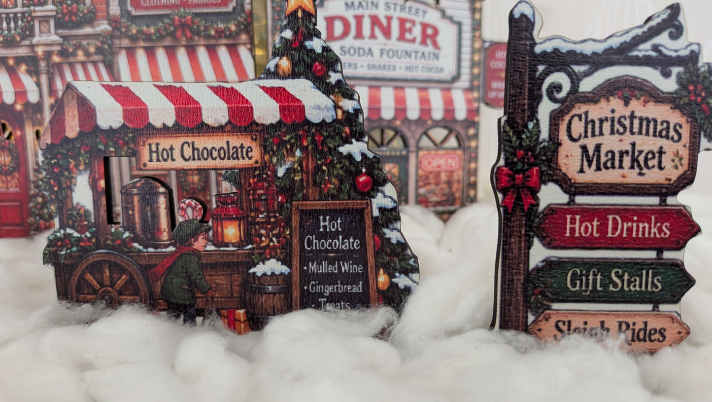 A decorative tabletop scene with a small hot chocolate stand, a Christmas tree, and a sign reading 'Christmas Market' with directional signs for hot drinks, gift stalls, and sleigh rides, set in front of a printed diner storefront background.