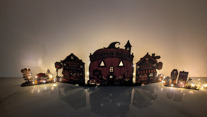 A Halloween-themed tabletop display featuring a central pumpkin-shaped house labeled 'Pumpkin Hollow' with smaller haunted house and tombstone decorations, lit by warm string lights against a dark background.