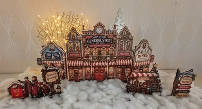 A decorative miniature village scene featuring a central general store with red-and-white striped awnings, side buildings including a diner and top shop, small figures, a red mailbox, and string lights in the background, arranged on white faux snow against a light patterned wall.